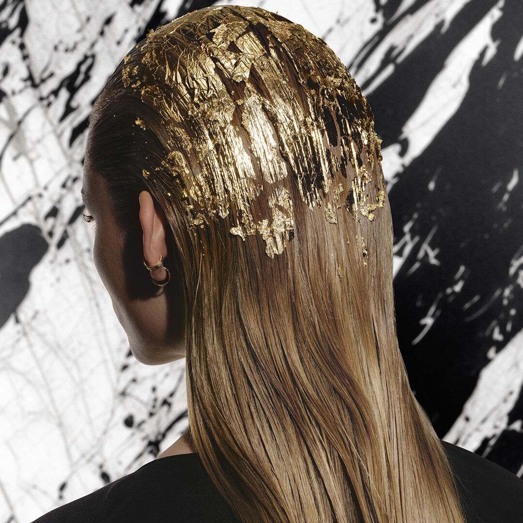 Donna bionda ripresa di spalle, con foglie d'oro tra i capelli e accappatoio nero Carita.