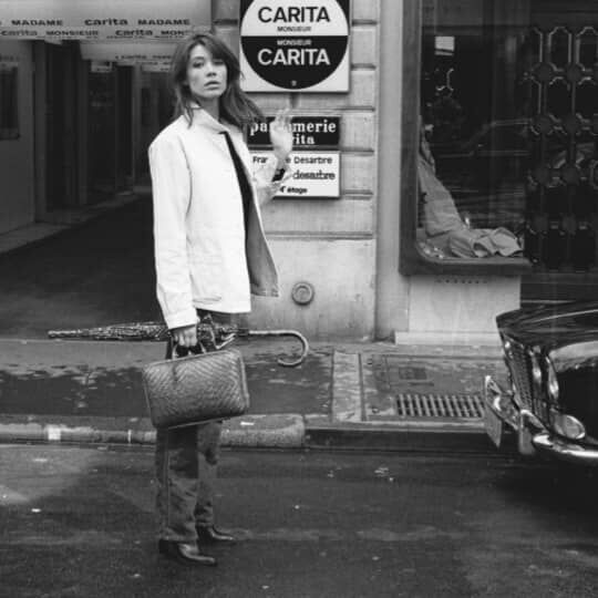 Una donna in bianco e nero in un vicolo, guardando verso la camera, con un cartello che riporta la scritta "CARITA" in vista.