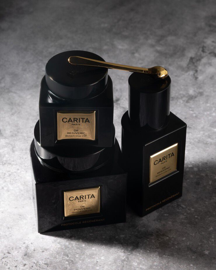 Una lussuosa esposizione di prodotti per la cura della pelle Carita Paris "Or Rejuvenic" e una spatola dorata, disposti elegantemente su una superficie scura e texturizzata, evidenziando un protocollo rigenerante.