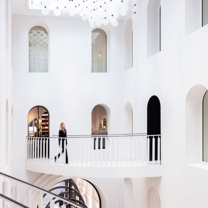 Carita Maison de Beaute: interni luminosi e moderni con pareti bianche, archi eleganti, balcone curvo con persona e soffitto con cerchi luminosi.