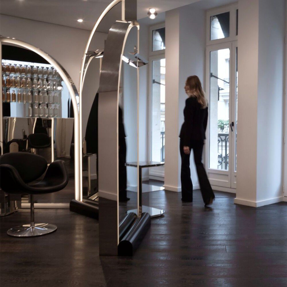 Un elegante e moderno salone da parrucchiere. Grandi strutture ad arco riflettenti con illuminazione integrata. Una raffinata sedia nera e visibile sulla sinistra. Scaffali a muro espongono bottiglie. Una donna cammina verso le finestre con un balcone parigino.