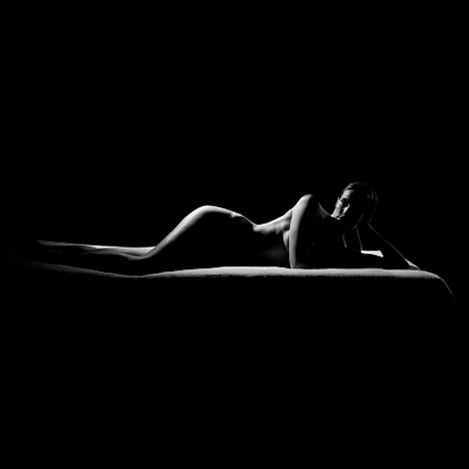 Una figura femminile nuda sdraiata su un lettino, illuminata da un'illuminazione soft che crea un forte contrasto con lo sfondo scuro.