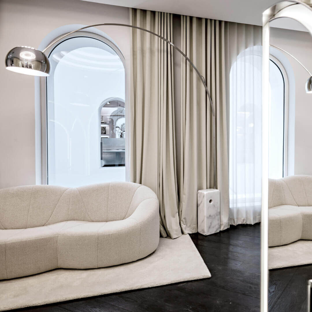 Moderna area lounge. Un divano beige dalla forma organica poggia su un tappeto abbinato, di fronte a finestre ad arco con tende beige. Una lampada ad arco lo illumina. Uno specchio verticale riflette la scena, aggiungendo spaziosita e luminosita.