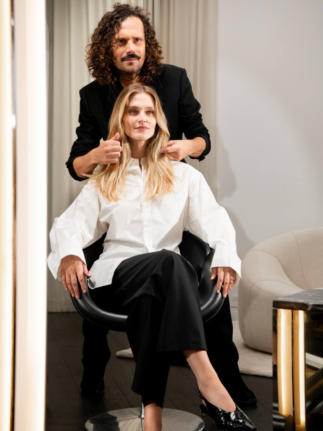 Un maestro hairstylist, con un gesto sicuro, perfeziona i capelli biondi e luminosi di una cliente, incarnando il lusso su misura e l’eccellenza artigianale del salone.