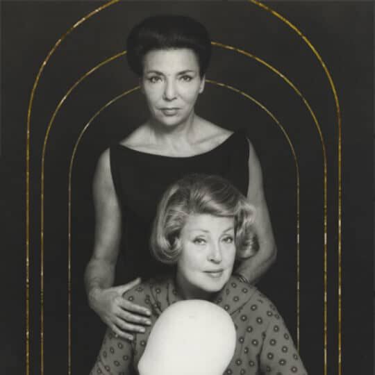 Due donne in abiti eleganti si trovano in un'illustrazione in bianco e nero con un fondo scuro. Una donna e in piedi dietro l'altra, che e seduta e tiene un oggetto circolare.