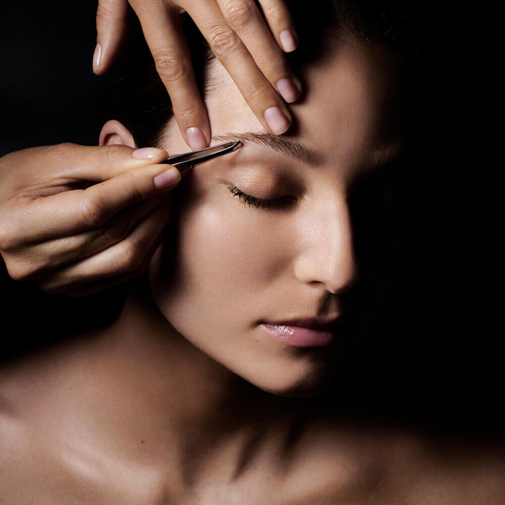 Primo piano del volto di una persona con un occhio chiuso, mentre una mano usa delle pinzette per modellare le sopracciglia su uno sfondo scuro.