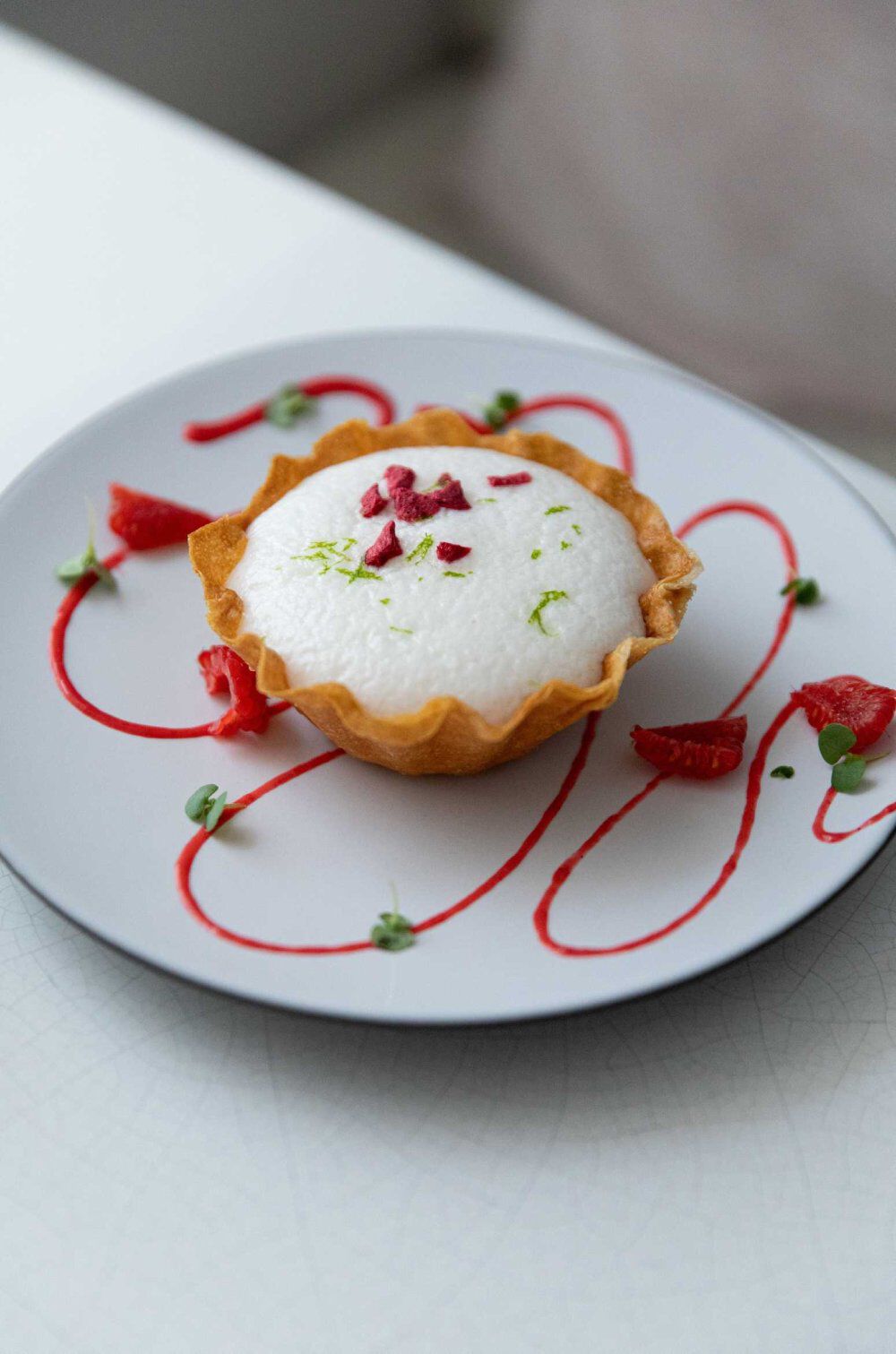 A dessert tart filled with a creamy, light-colored mousse, garnished with lime zest and small pieces of red fruit, sits on a white plate decorated with a swirl of red sauce and green microgreens.