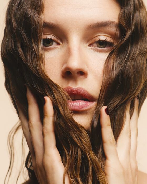 Primo piano di una donna con capelli castani mossi, che incornicia il viso con le mani su uno sfondo beige chiaro.