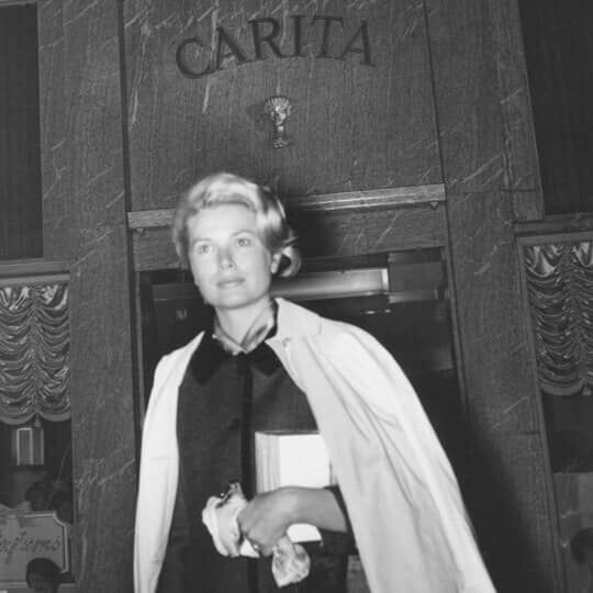 Una donna bionda con un'espressione seria, in un ambiente elegante e vintage.