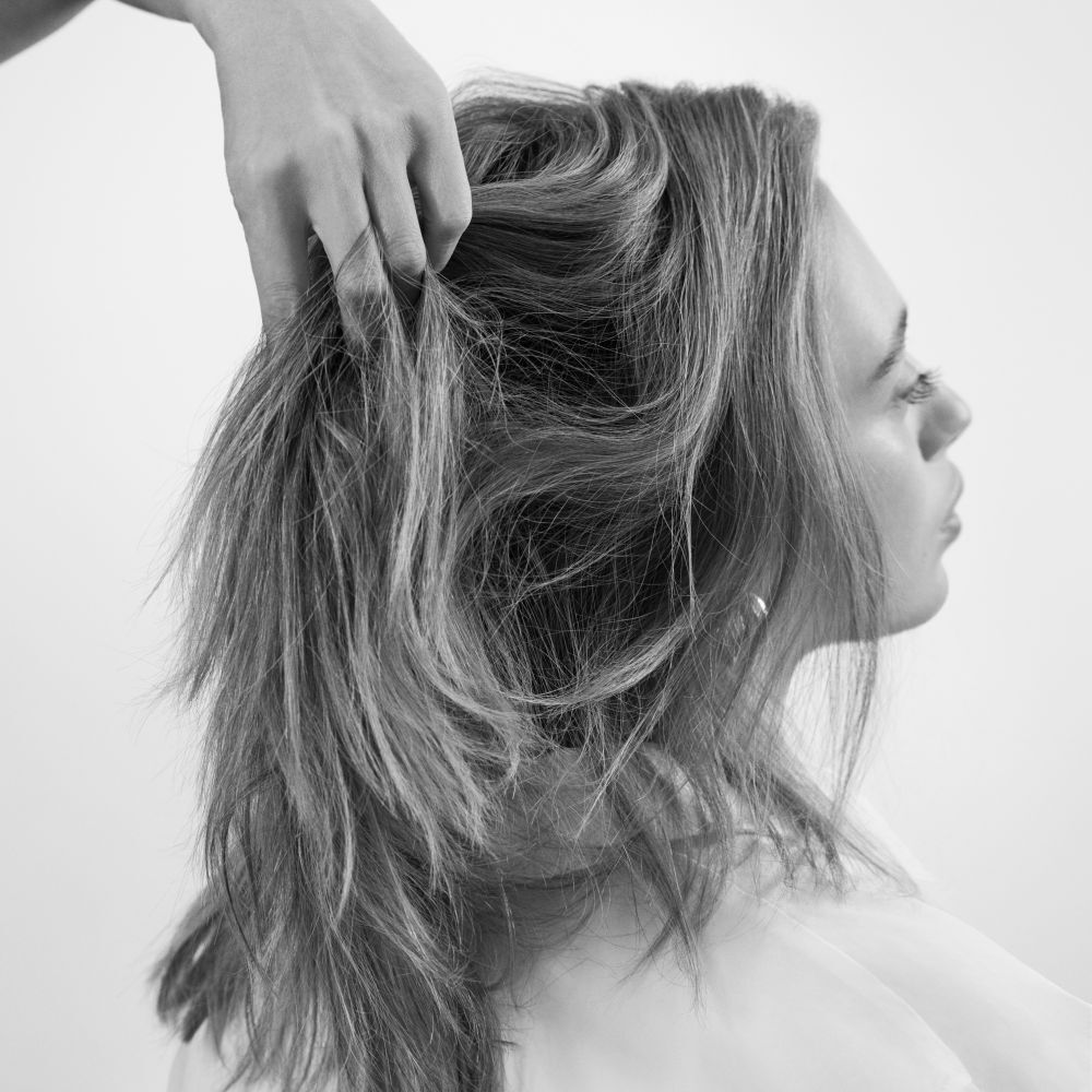 Un'immagine in bianco e nero mostra il profilo della testa di una persona, mentre una mano solleva delicatamente i suoi capelli di media lunghezza, ondulati e voluminosi, evidenziandone la texture e il movimento.