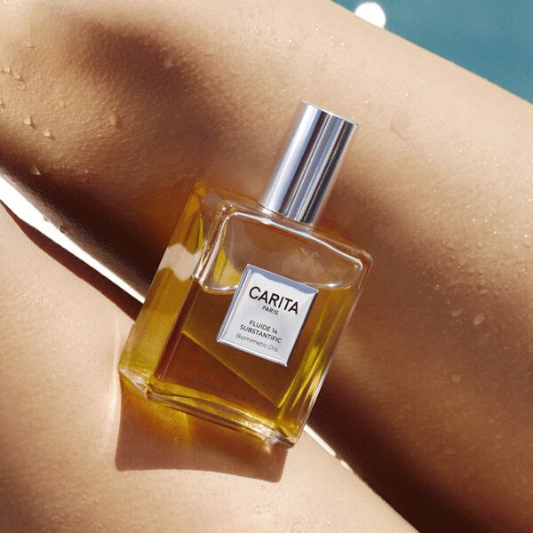 Un flacone di vetro dell'olio secco Fluide 14 di Carita adagiato su una pelle dorata dal sole e bagnata da gocce d'acqua, con il riflesso azzurro di una piscina sullo sfondo.​