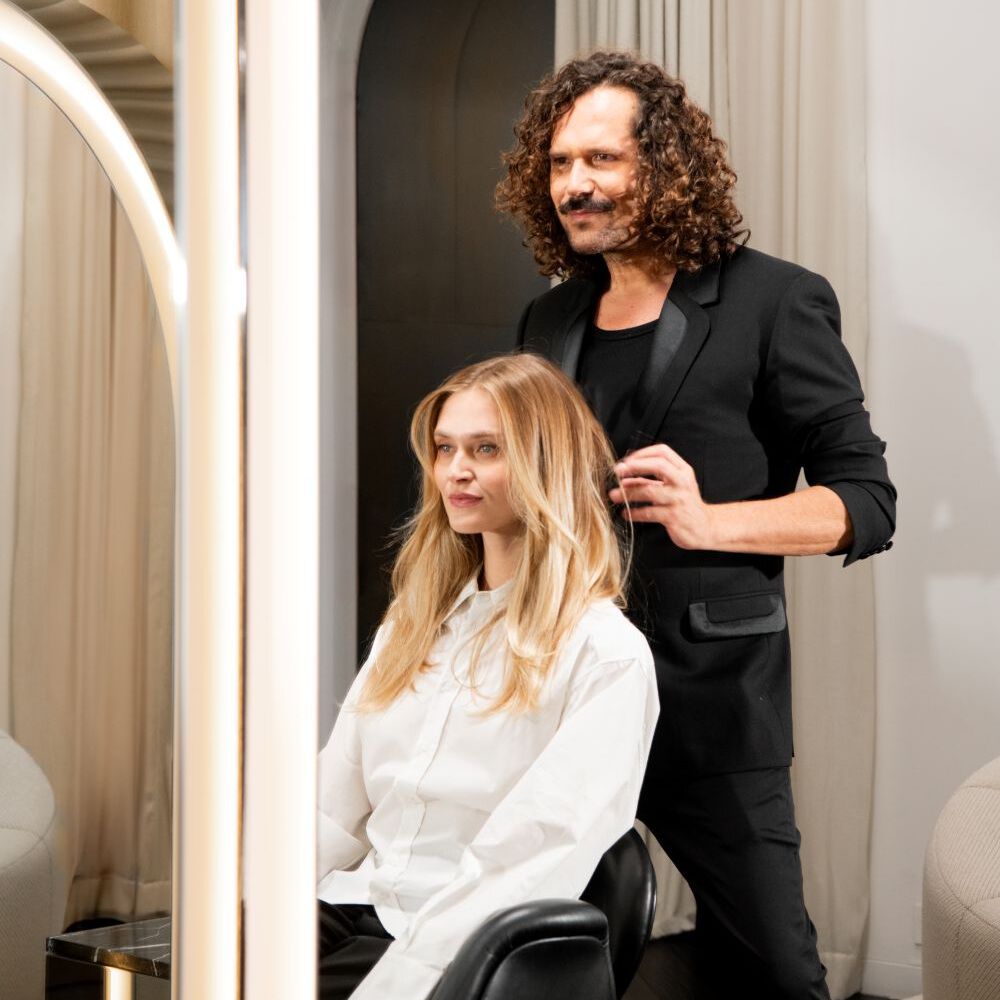 In un salone moderno, una donna bionda in camicia bianca siede su una sedia nera, guardando a sinistra. Un uomo in abito nero le sistema i capelli. La scena e incorniciata da specchi che riflettono tende beige chiare e una scura porta ad arco.