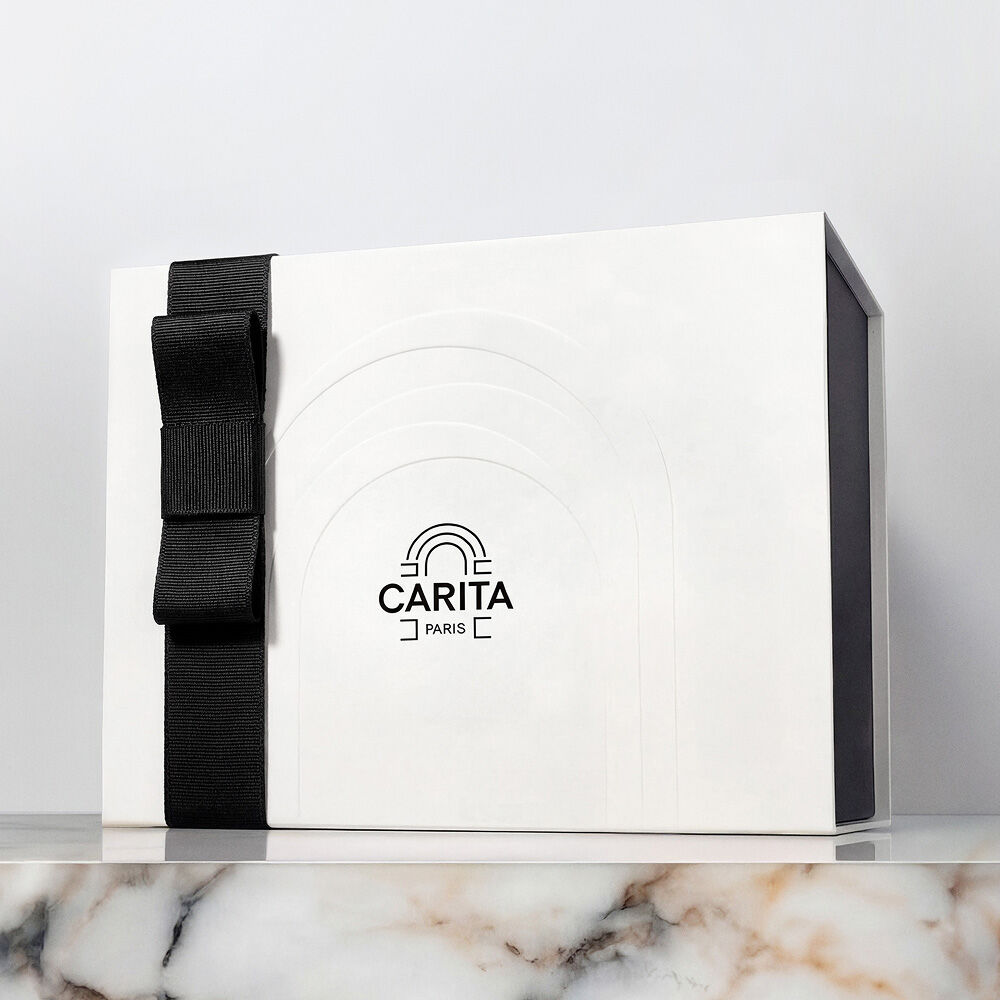 Una confezione regalo di lusso bianca Carita Paris con un elegante fiocco di nastro nero, presentata su una superficie di marmo.