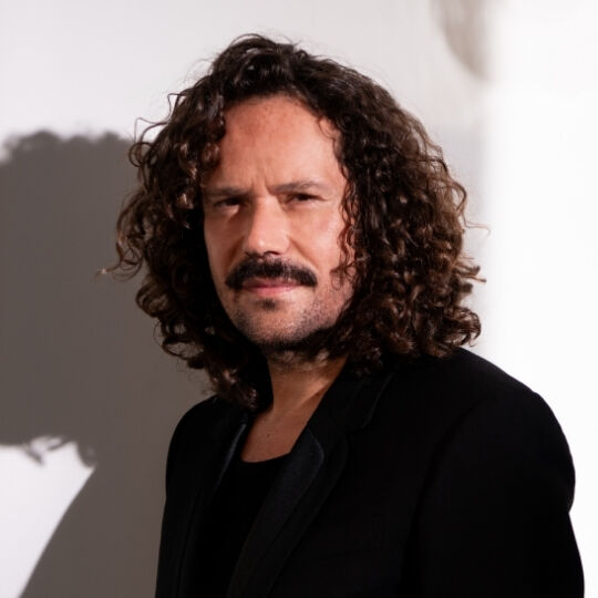 Ritratto di un uomo che guarda direttamente, con baffi prominenti e capelli folti e ricci.