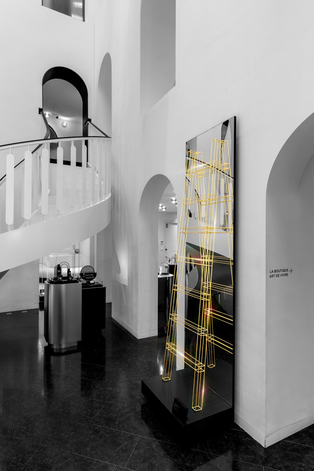 A modern interior featuring a staircase and a vertical display with yellow line art resembling a ladder. The space has minimalist decor with white walls and dark flooring.