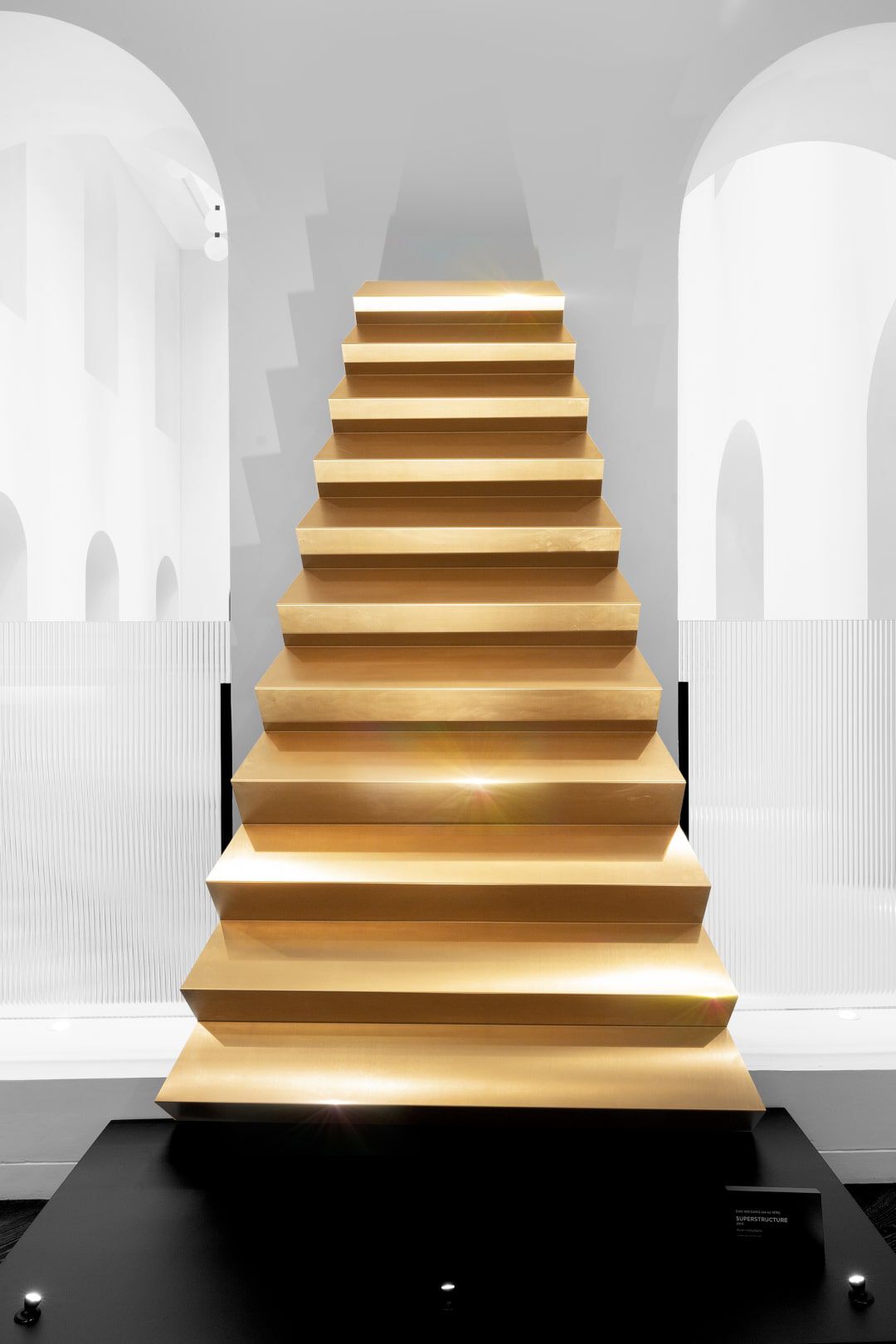 A minimalist, golden staircase with shallow steps, set against a white background with arched openings.