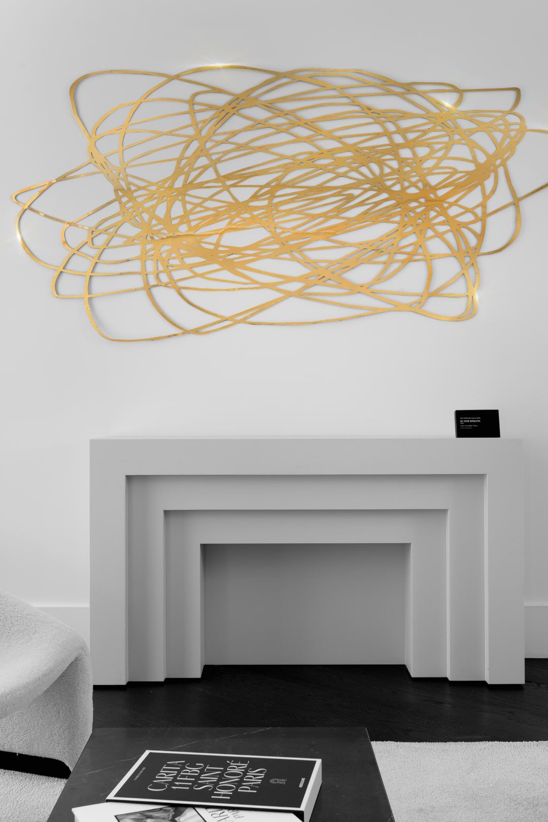 A contemporary living space featuring a minimalist white console table with a geometric gold wall sculpture above it. A stack of books sits on the table, with a small black sign nearby. The walls are painted white, and the floor is dark.