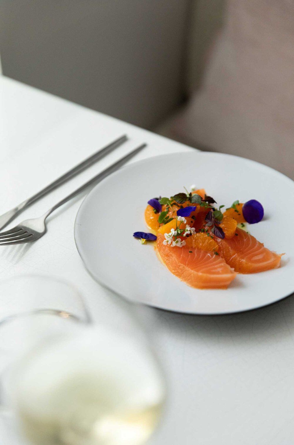 A white plate featuring sliced salmon topped with edible flowers and orange segments, accompanied by a fork and knife on a table. A glass of white wine is blurred in the foreground.