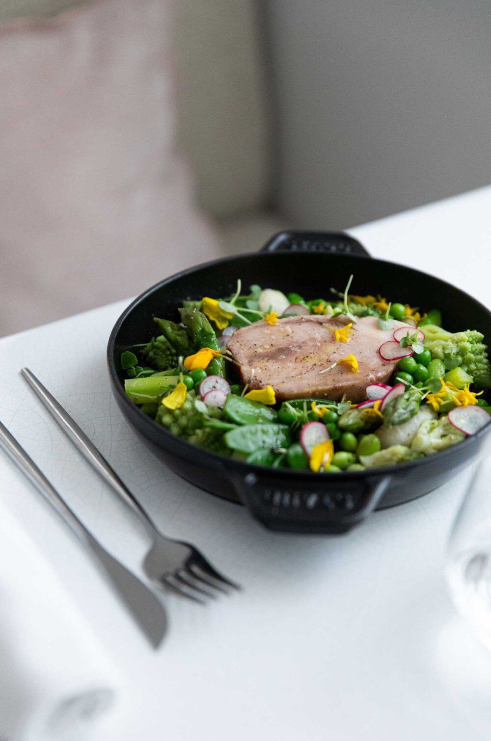 A vibrant salad composed of various greens, radishes, and edible flowers, topped with a piece of sauteed fish, arranged in a black skillet on a table setting with utensils.