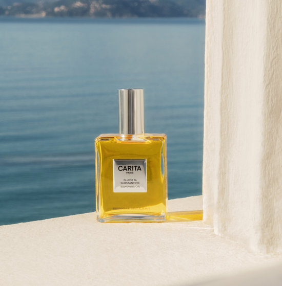 Un flacone dell’olio secco Fluide 14 nel suo formato quadrato, con tappo e targhetta superiore.​ E posizionato sul davanzale di una finestra senza vetro, con le montagne e il mare sullo sfondo, evocando l’estate.​