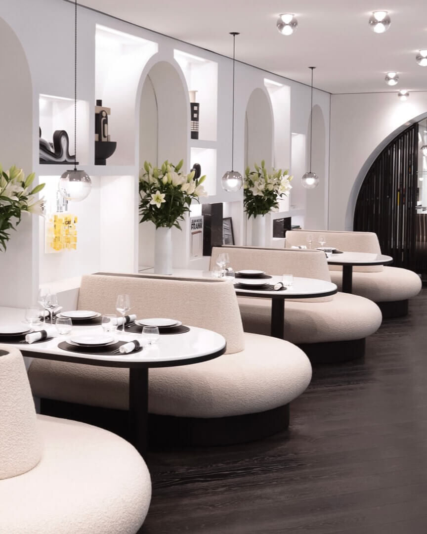 Elegante sala da pranzo con panche curve, tavoli rotondi apparecchiati, pareti bianche arcuate con nicchie e bouquet di gigli bianchi. Ambiente chic e moderno.