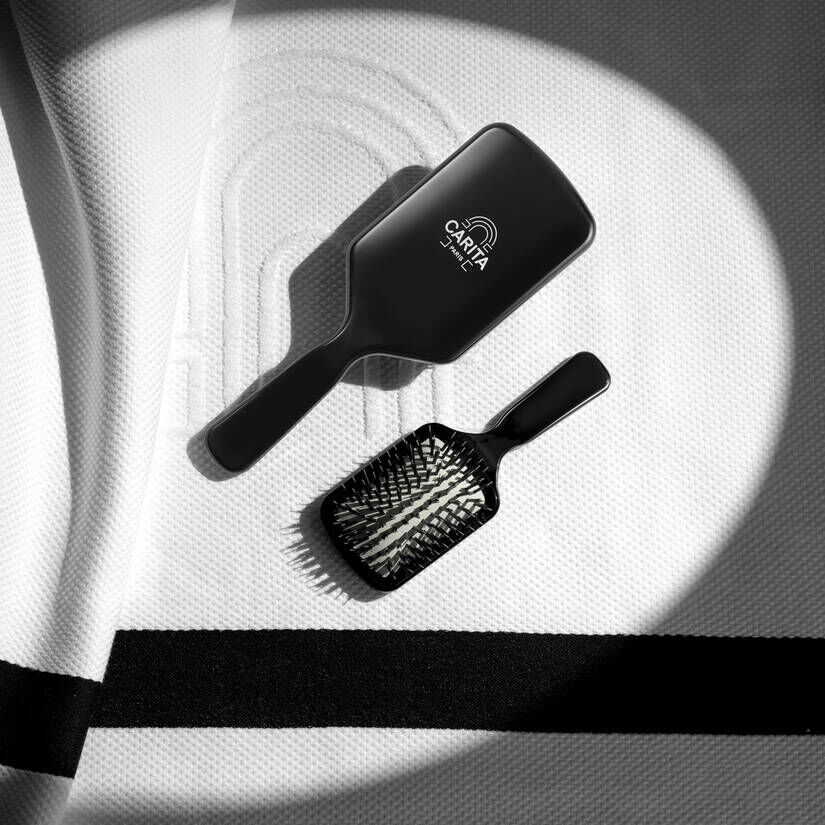 Due spazzole nere per capelli, una grande e una piu piccola, sono adagiate su una superficie bianca testurizzata, forse un panno di spugna o una superficie uniforme. La spazzola piu grande, situata in alto a sinistra, mostra il logo CARITA Paris con il motivo ad arco. La spazzola piu piccola e posizionata in basso a destra. Ombre nette sono visibili sulla superficie.