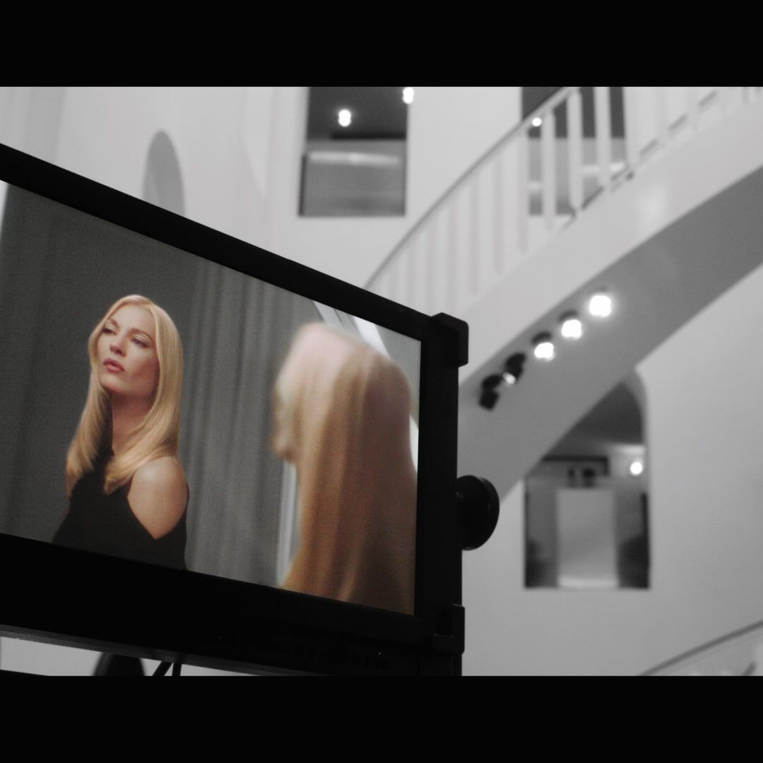 A black and white image featuring a monitor displaying a model with long blonde hair looking into a mirror, set against a backdrop of a modern, minimalist interior with a staircase and arches.
