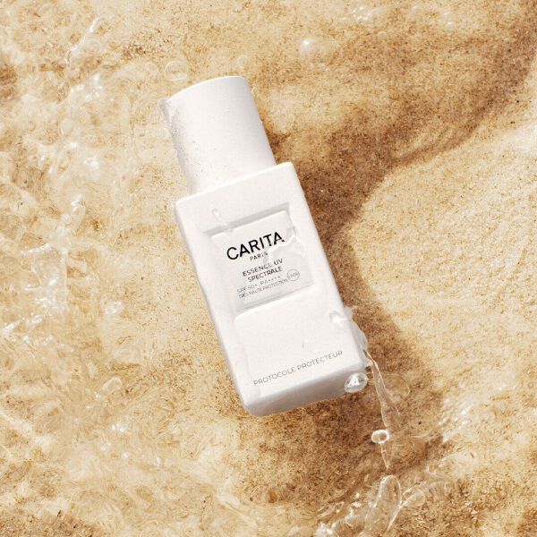 Flacone bianco dell'Essence UV di Carita appoggiato sulla sabbia.