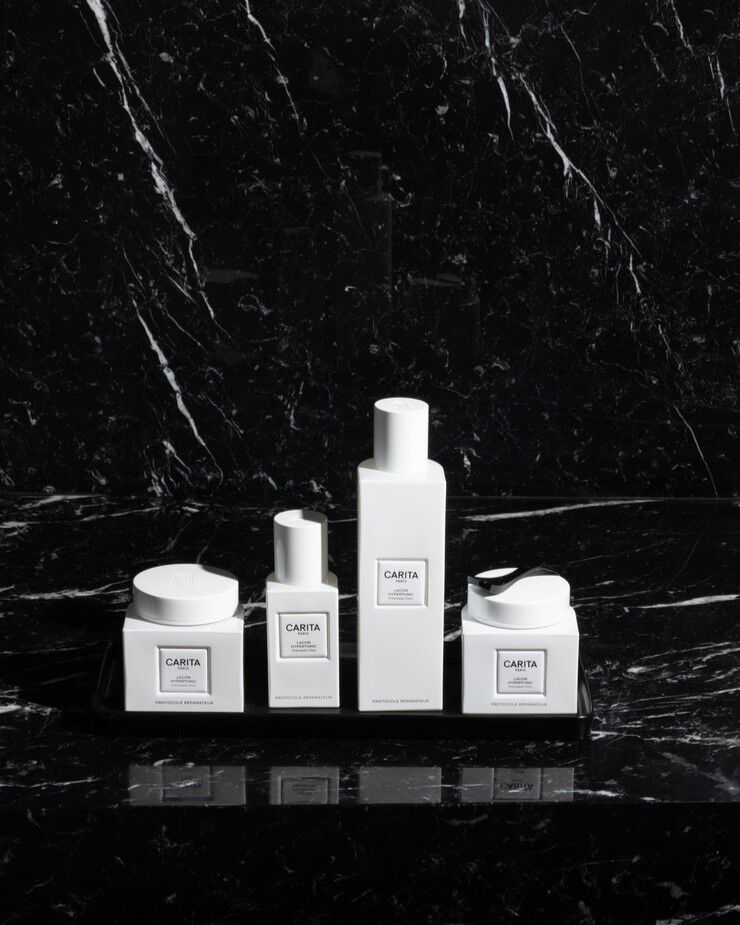 Un'elegante collezione di prodotti per la cura della pelle Carita Paris "Lagon Hypertonic" bianchi, inclusi vasetti e flaconi, esposti su una raffinata superficie in marmo nero, evidenziando lusso e minimalismo.