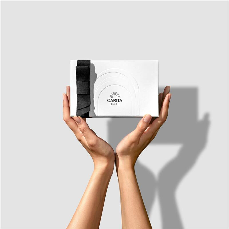 Mani di una donna che tengono una confezione regalo di lusso bianca Carita Paris, decorata con un elegante fiocco di nastro nero.