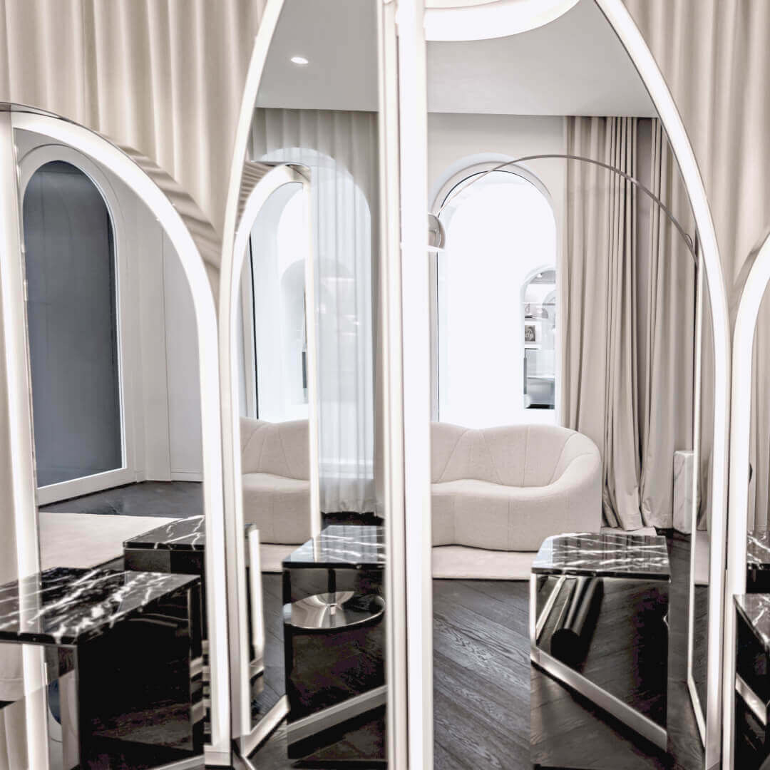 Questo elegante interno moderno presenta specchi riflettenti ad arco con illuminazione lineare. Tavoli in marmo nero e un pavimento in legno scuro sono visibili. Un divano chiaro, tende beige e una lampada ad arco creano un'atmosfera raffinata.