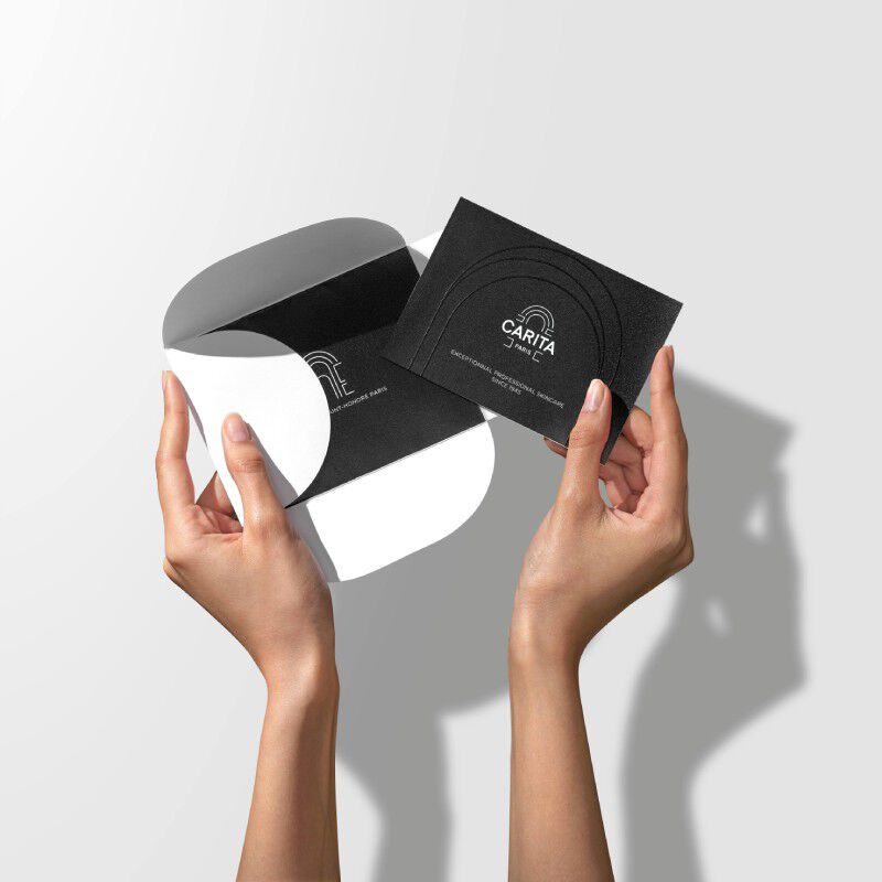 Mani che tengono una card Carita Paris nera minimalista e una busta grafica in bianco e nero su uno sfondo neutro.