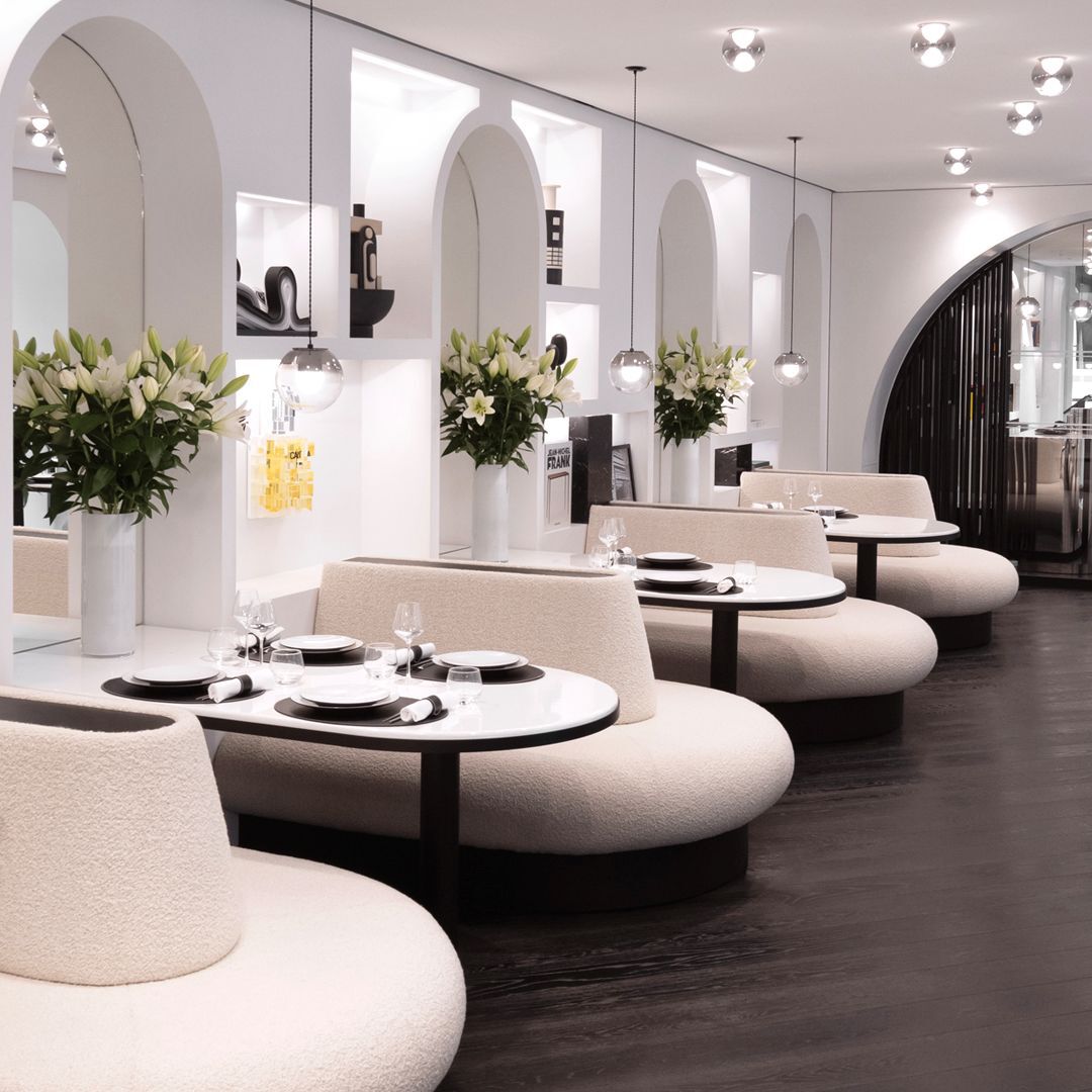 Elegante sala da pranzo con divanetti curvi, tavoli rotondi, pareti bianche ad arco con nicchie e bouquet di gigli bianchi. Ambiente chic e moderno.