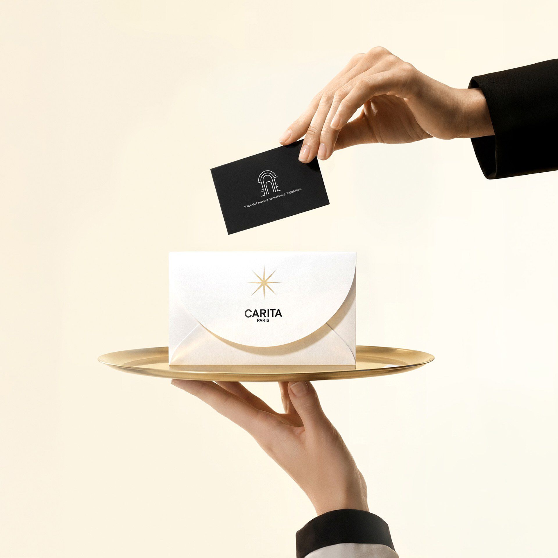 Una mano presenta una busta Carita su un vassoio dorato, mentre un'altra vi inserisce una carta regalo nera con il prestigioso indirizzo di 11 Faubourg Saint-Honore.