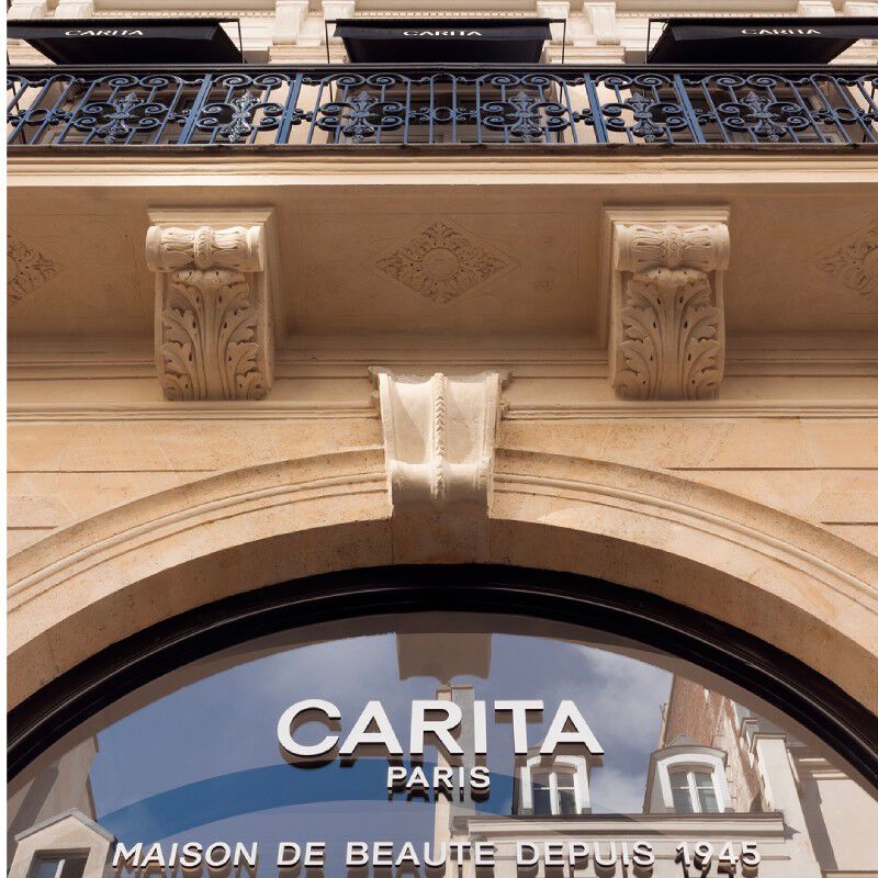 Elegante facciata della Maison Carita Paris. Si vedono un balcone in ferro battuto, dettagli architettonici scolpiti e il logo Carita Paris sulla vetrina curva. Marchio "Maison de Beaute Depuis 1945".